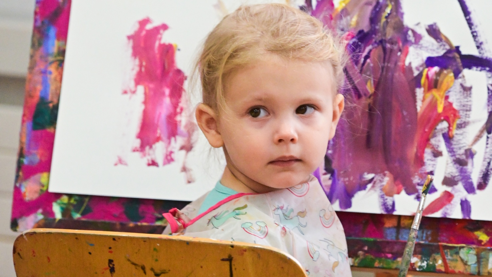 Kleinkind mit Malschürze sitzt vor bunten Kindermalereien in einem Kurs der Kunstschule Liechtenstein.
