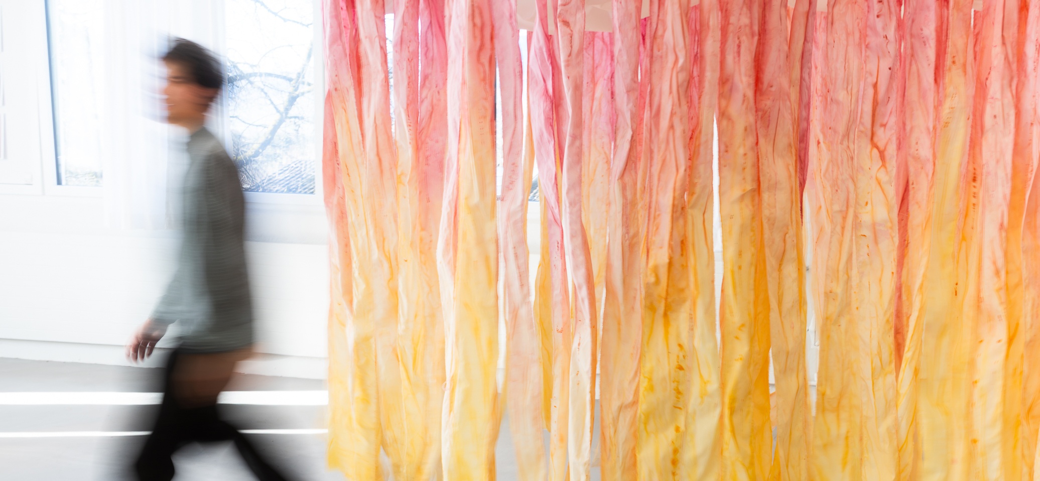 Person geht an einer raumgrossen Textilinstallation aus hängenden, gelb-orange-rosa gefärbten Stoffstreifen vorbei.