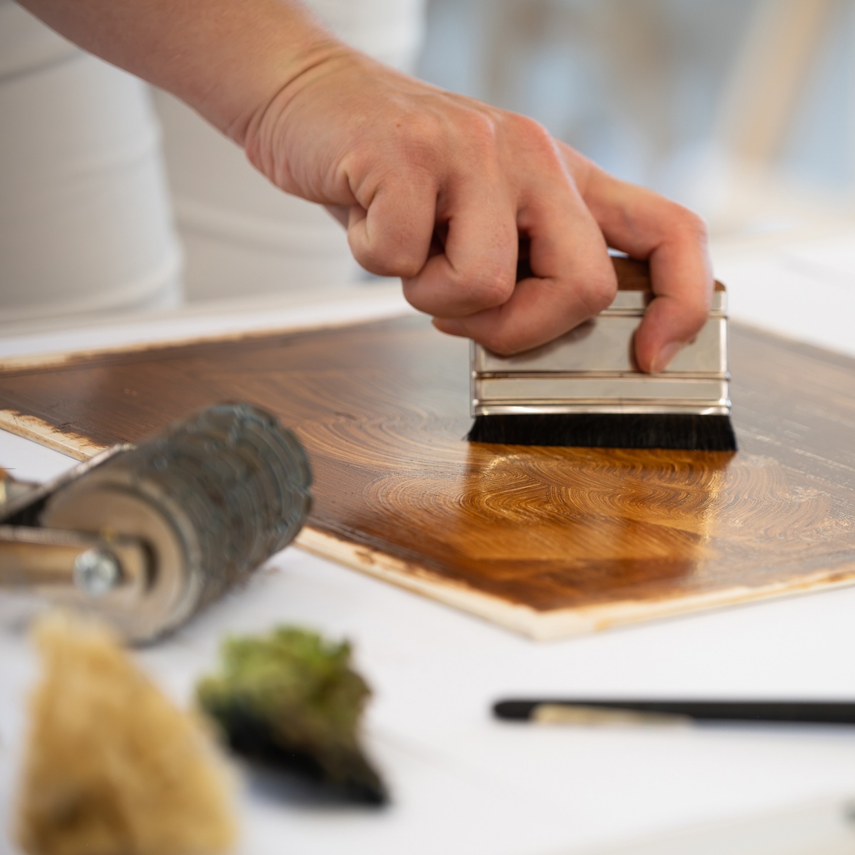 Hand trägt mit einem Borstenpinsel Beize auf eine Holzplatte auf.